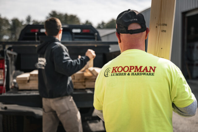 Two men loading wooden planks into a truck with one wearing a yellow Koopman Lumber & Hardware shirt.