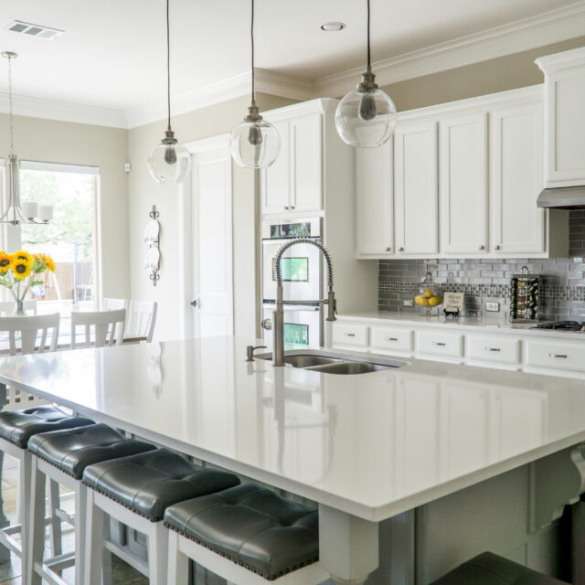 Modern kitchen with white cabinets, large island, pendant lights, and stools. Sunflowers on the dining table in the background.