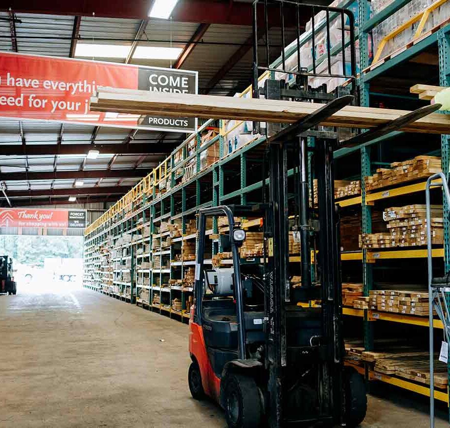 Forklift lifting wooden planks in a warehouse aisle with shelves stocked with building materials and supplies.