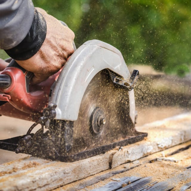 Person using a circular saw to cut wood, with sawdust flying, in an outdoor setting.