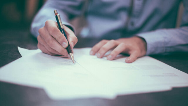 Person writing with pen on paper at a desk, wearing a blue shirt, focused on work.