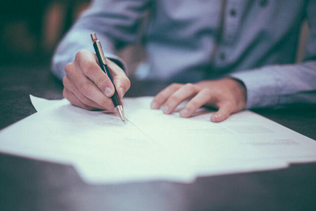 Person writing with pen on paper at a desk, wearing a blue shirt, focused on work.