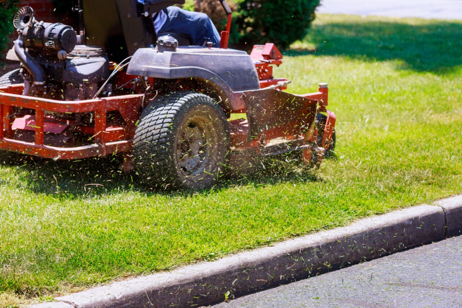 Riding lawn mower cutting bright green grass, with clippings flying in sunny outdoor garden setting.