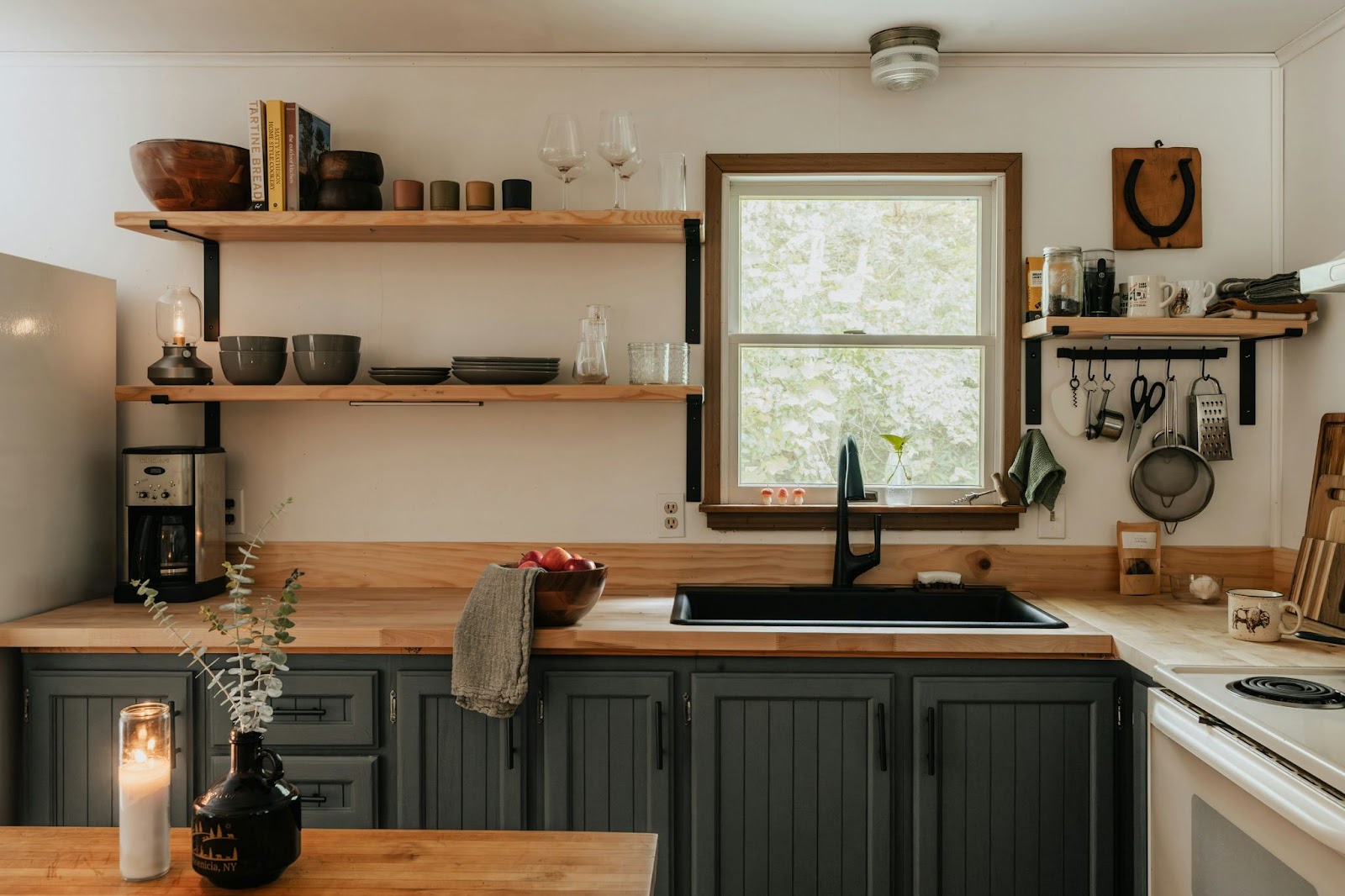 A cozy kitchen uses open wood shelving near the stove and sink to store kitchen utensils, ceramic dinnerware, and cookbooks.