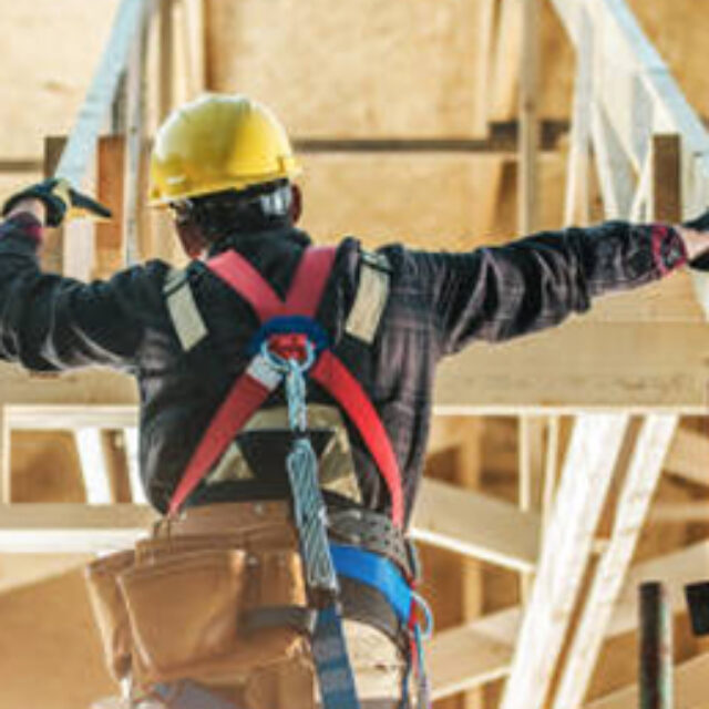 Construction worker wearing safety gear and a helmet, balancing on a wooden structure at a building site.