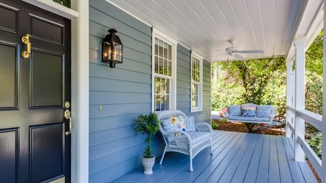A blue house has a black door with brass hardware and a large porch with a white chair, a porch swing, a ceiling fan and a potted plant.