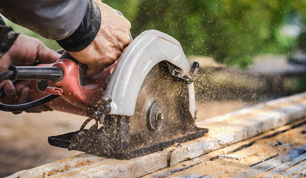 Person using a circular saw to cut wood with sawdust flying around in an outdoor setting.