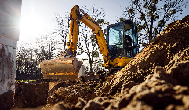 Yellow excavator digging soil at a construction site with bare trees in the background on a sunny day.