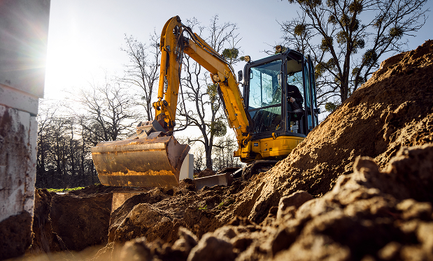 Yellow excavator digging soil at a construction site with bare trees in the background on a sunny day.