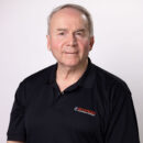 Older man in a black polo shirt standing against a plain white background.