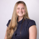 Smiling person with long hair wearing a dark polka dot blouse against a plain background.