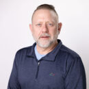 Man with short hair and beard wearing a blue collared shirt against a plain white background