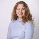 Smiling person with curly hair wearing a light blue sweater against a plain white background.