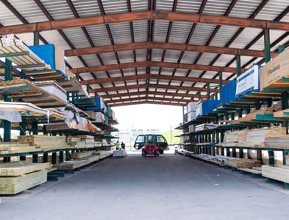 Covered lumber storage area with two vehicles parked between rows of stacked wood material.