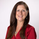 Smiling person with long brown hair wearing a red blouse, standing against a plain white background.