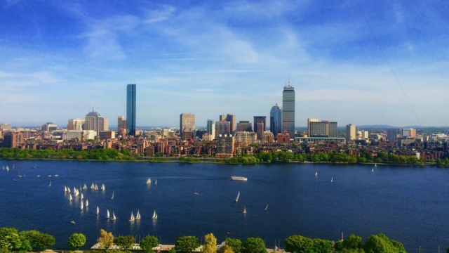 The picture depicts The Boston skyline with skyscrapers, buildings, and a body of water. The body of water has sailboats and connects to a dock across from the cityscape.