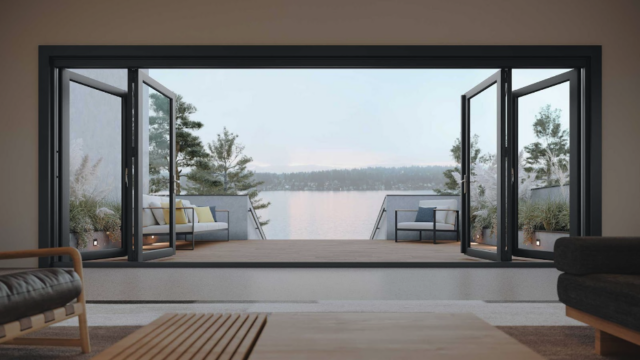 The picture depicts an open glass door that overlooks a body of water and a forest on the coastline.