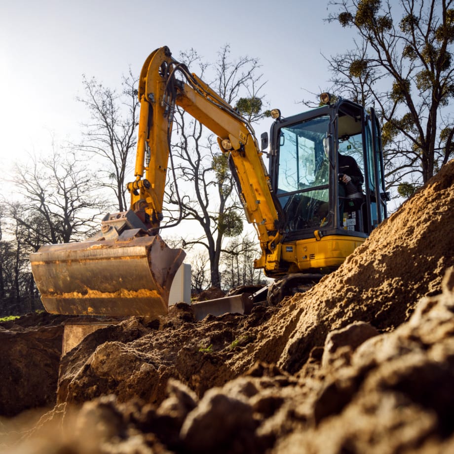 Yellow excavator digging large mound of earth at a construction site with trees in the background.