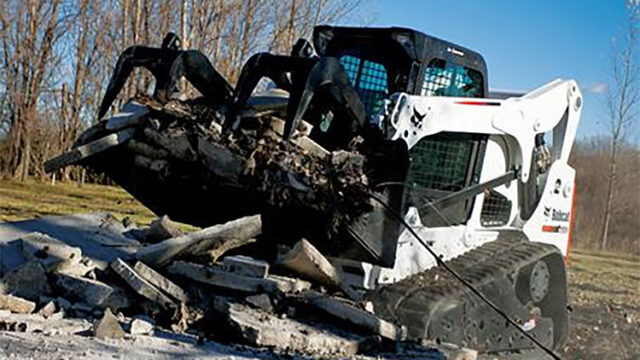 Skid steer loader with grapple attachment clearing debris on a sunny day, surrounded by trees in the background.