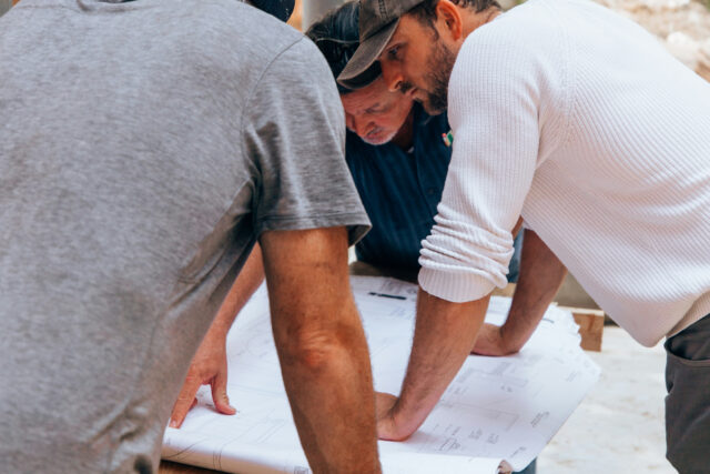 Three people reviewing blueprints at a construction site, focusing intently on plans, wearing casual work attire.