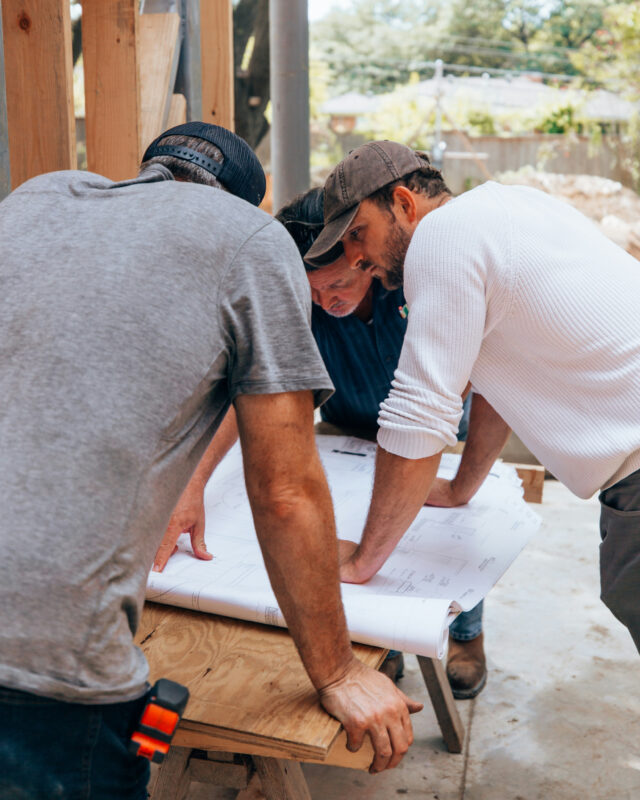 Three people reviewing blueprints at a construction site, focusing intently on plans, wearing casual work attire.