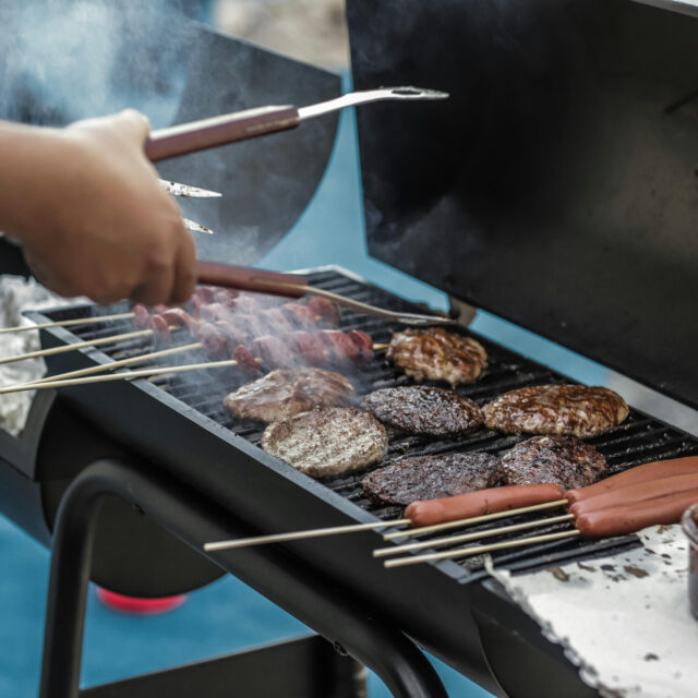 Person grilling burgers and hot dogs on a charcoal grill with smoke rising.