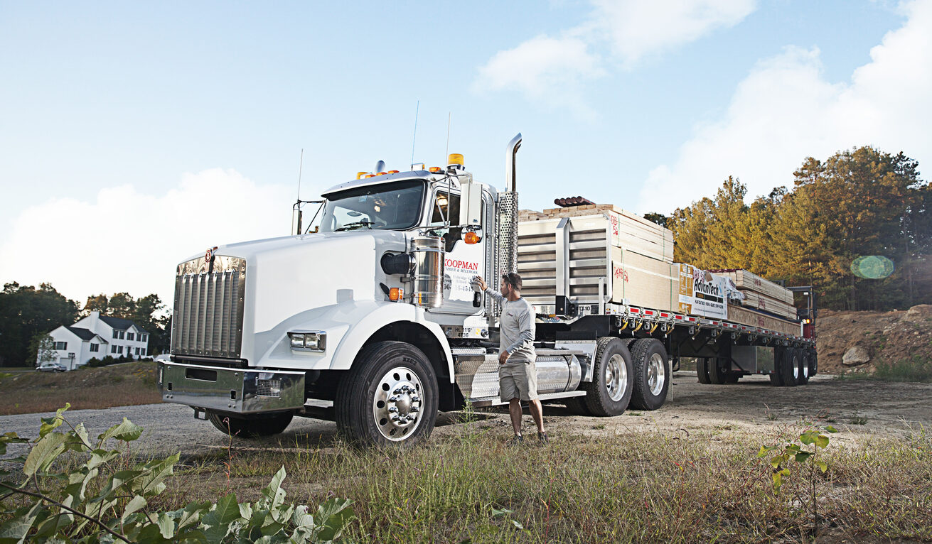 Large white semi-truck parked outdoors with a person standing beside it on a grassy area under a partly cloudy sky.