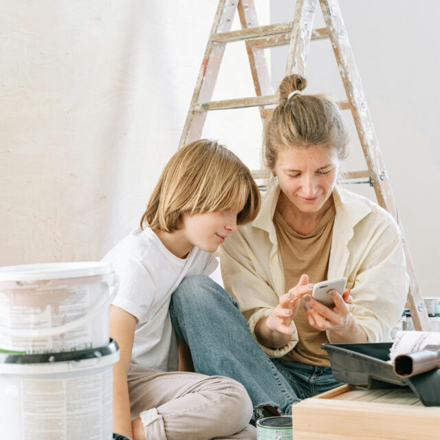 Two people seated on the floor, discussing paint choices in a room with painting supplies and a stepladder in the background.