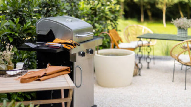 Outdoor patio with a gas grill, table, and chairs surrounded by greenery.
