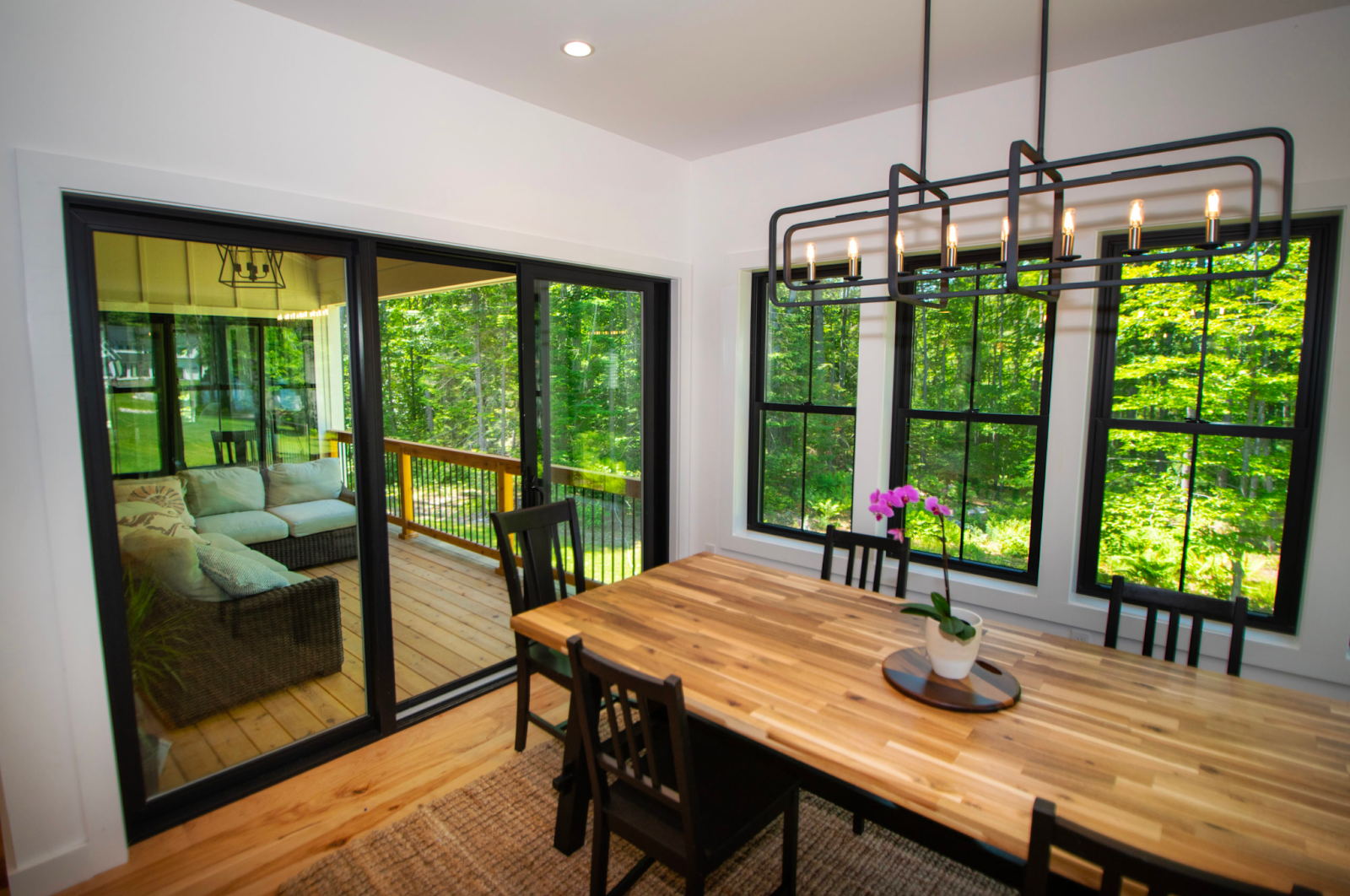 The picture depicts a dining room with a wooden table, decorated with a potted orchid. Three black double-hung windows sit behind the table, and a glass sliding door leads to a covered deck.