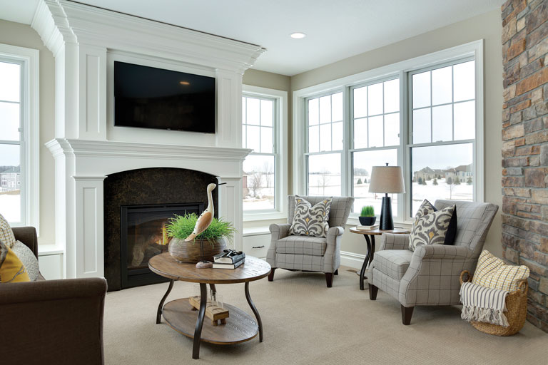 The picture depicts a living room with a TV mounted above a fireplace and two chairs adjacent to the fireplace. There is a wooden coffee table with a bird statue on it and windows on both sides of the walls.