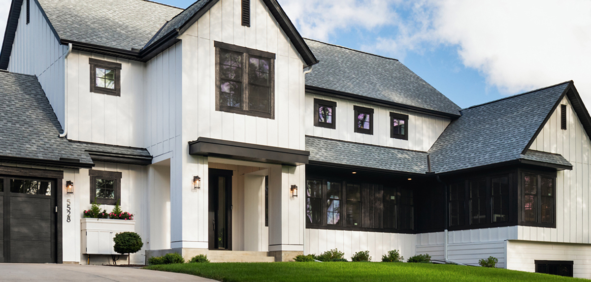 The picture depicts a large modern-style farmhouse painted white with slate roofing. There is a covered entryway and windows with black paneling. 