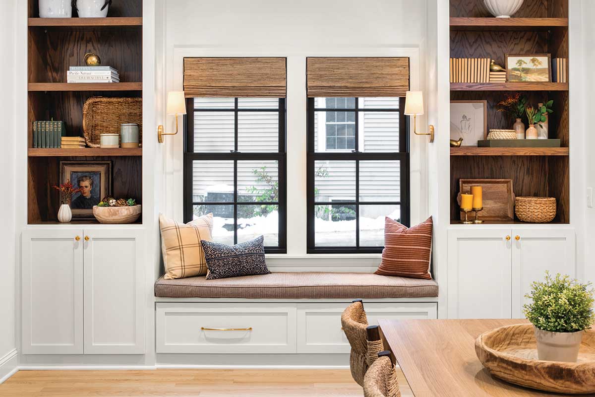 The picture depicts a dining room wall with built-in cupboards and a built-in sofa joining the cupboards together. The cupboards are filled with knick knacks and the sofa has two large glass windows above it. 