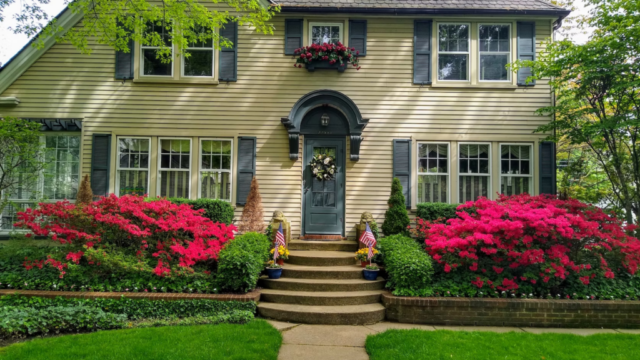 Two-story house with arched entryway, surrounded by vibrant pink azaleas and lush green lawn, under a clear sky.