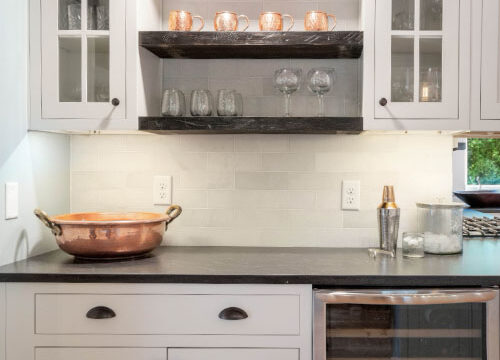 Modern kitchen with white cabinets, dark countertop, copper accents, and wine fridge. Shelves hold glassware and decor.