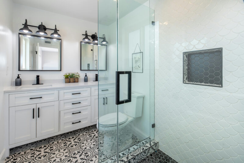 Modern bathroom with glass shower, double vanity, decorative tile floor, and black fixtures.