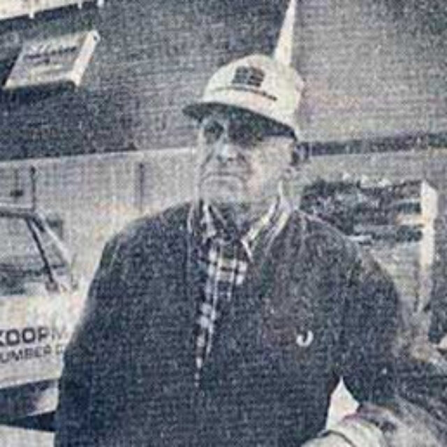 Man wearing a cap and jacket stands in front of a building with a sign, looking away from the camera.