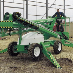 Man operating a green specialized lift with articulated arms and wheels inside a large industrial facility.