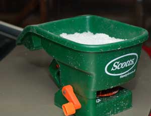 Green garden spreader with white powder inside, handle and orange adjustment lever visible on concrete surface.