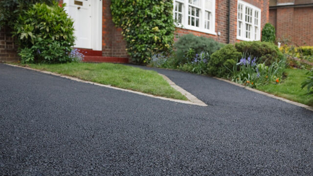 Freshly paved asphalt driveway leading to a brick house with neatly trimmed grass and shrubs on either side.