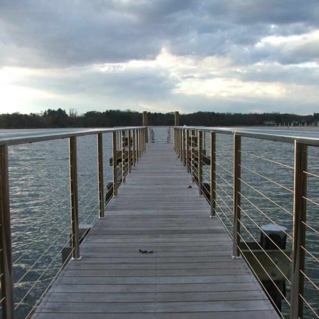 Wooden pier with metal railings extending over a calm body of water under a cloudy sky.