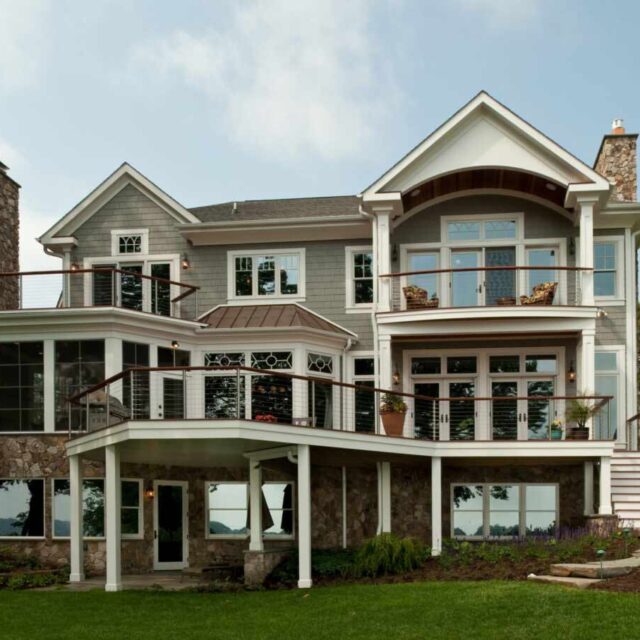 Spacious two-story house with large windows, wraparound porch, and lush green lawn on a clear day.