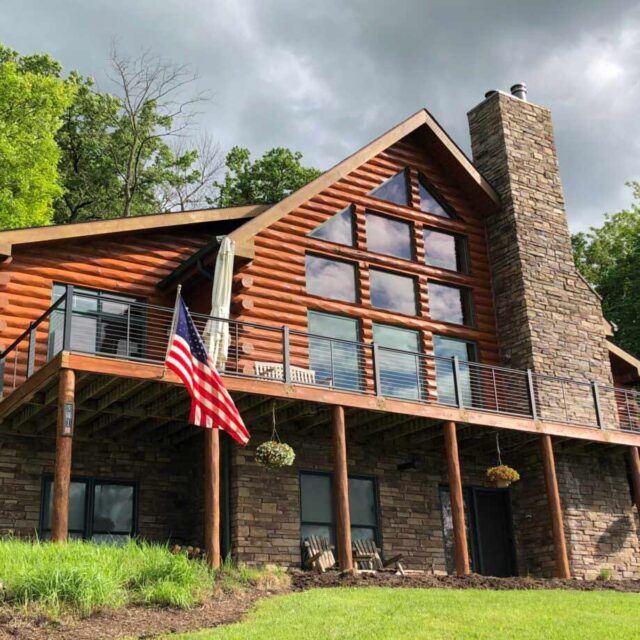 Rustic log cabin with large windows and stone chimney, surrounded by greenery, with American flag in front.