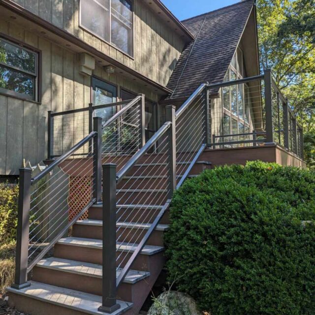 Modern house with wooden deck and cable railing, surrounded by greenery on a sunny day.