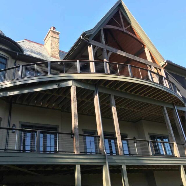 Modern two-story house with expansive curved balcony and wooden beams against clear blue sky.
