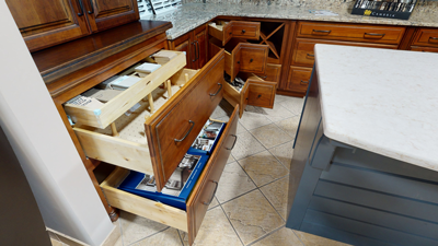 Open wooden kitchen drawers with visible contents like books and utensils in a tiled floor setting next to a kitchen island.