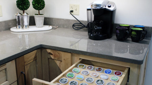 Kitchen counter with Keurig coffee maker, three mugs, a plant, and an open drawer showing assorted coffee pods.