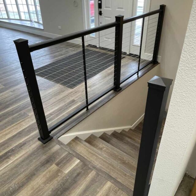 Black metal railing above a small staircase in a modern home with wood flooring and large windows in the background.