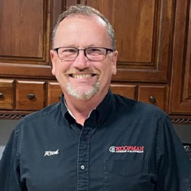 Smiling person wearing a dark shirt in a wooden cabinet kitchen setting.