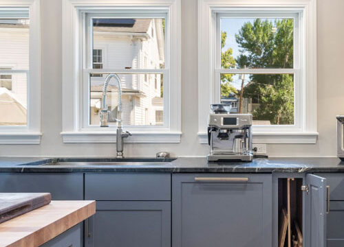 Modern kitchen with blue cabinets, black countertop, and coffee machine, illuminated by natural light through three windows.